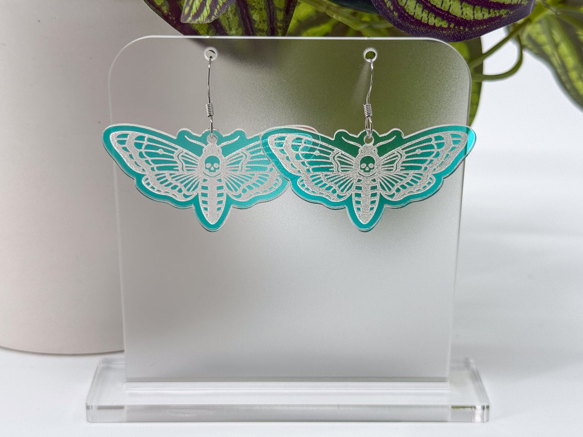 A pair of dangling earrings shaped like moths, made from iridescent acrylic, hanging from silver hooks on a clear acrylic display stand.