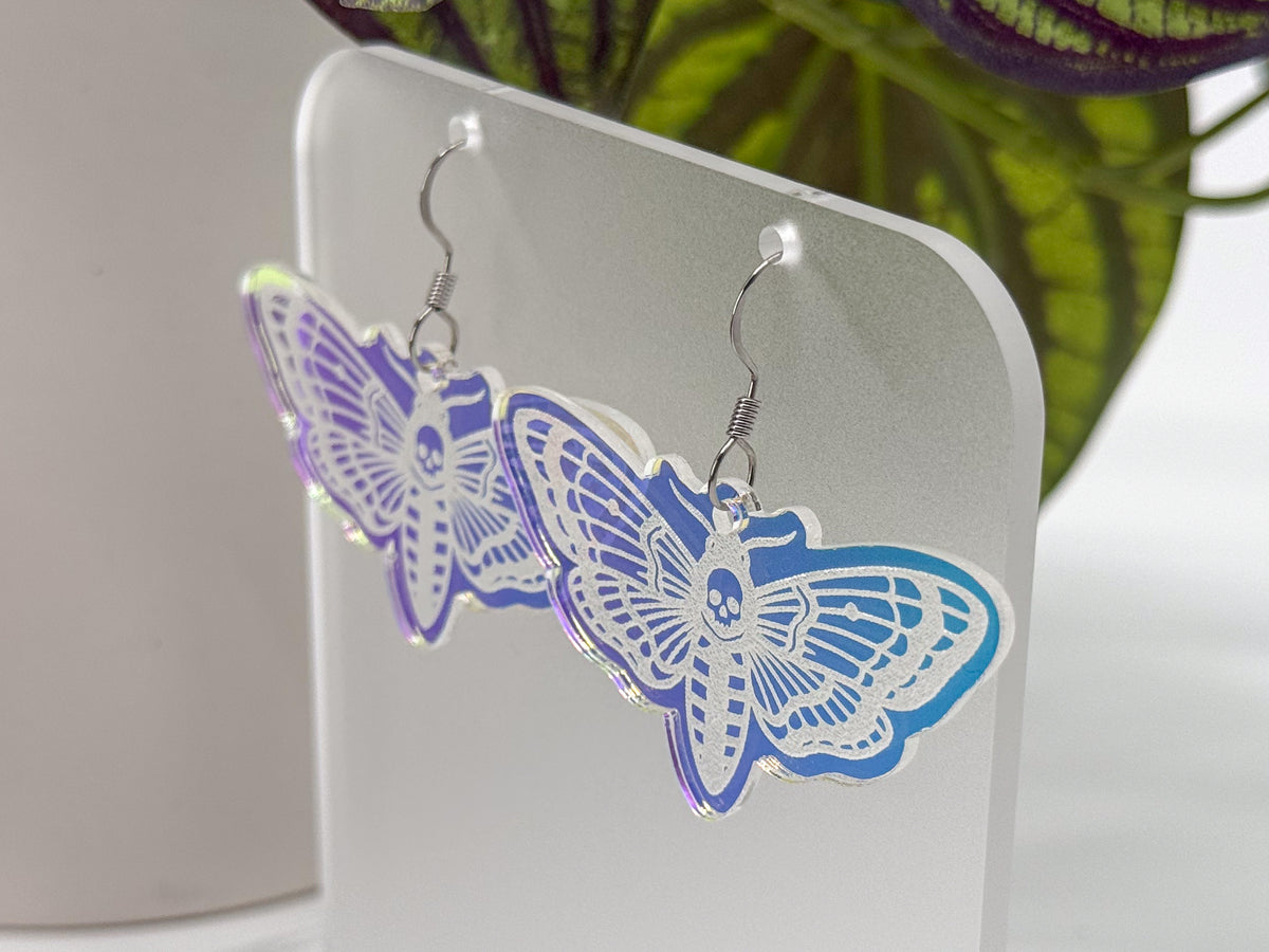 A pair of dangling moth earrings with white cutout butterfly designs and a purple-to-blue iridescent border, displayed on a clear acrylic stand.