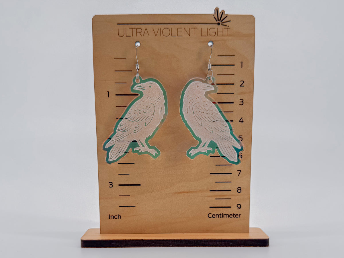 A pair of raven-shaped dangling earrings displayed on a wooden measurement stand labeled 'ULTRA VIOLENT LIGHT.' The earrings have a pale pinkish-beige center with a translucent iridescent outline, shown against a light gray studio background.