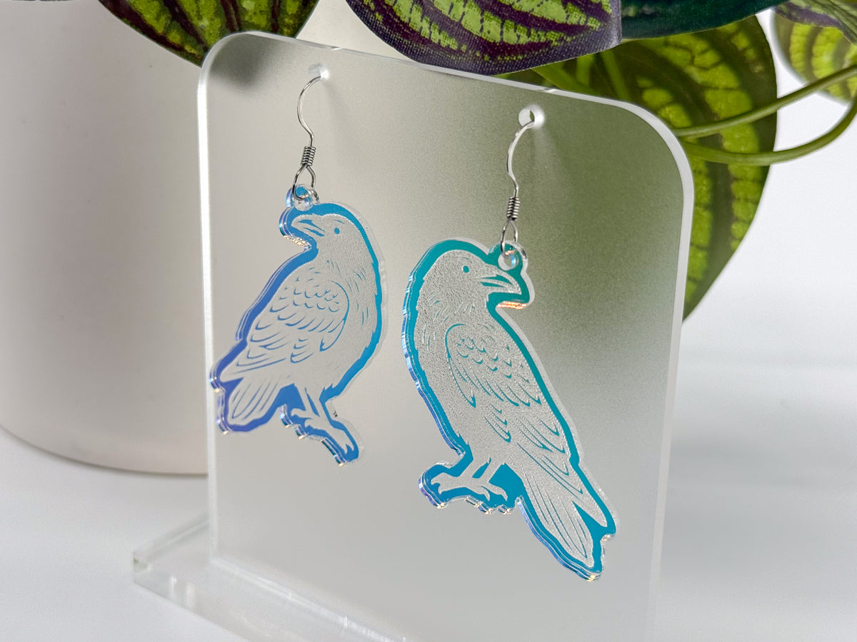 A pair of engraved iridescent raven-shaped drop earrings with silver hooks, displayed on a clear acrylic stand with a blurred white pot and green leaves in the background.