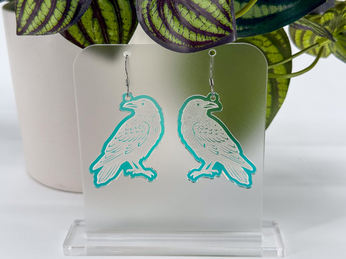 A pair of iridescent raven-shaped earrings displayed on a clear acrylic stand, with a white pot and green leaves in the background.