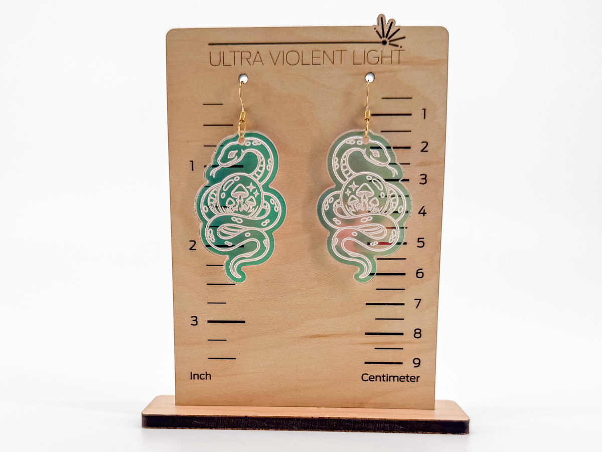 A pair of snake-shaped dangle earrings displayed on a wooden measurement stand labeled 'ULTRA VIOLENT LIGHT.' The earrings are iridescent acrylic with white snake patterns and gold-toned hooks.