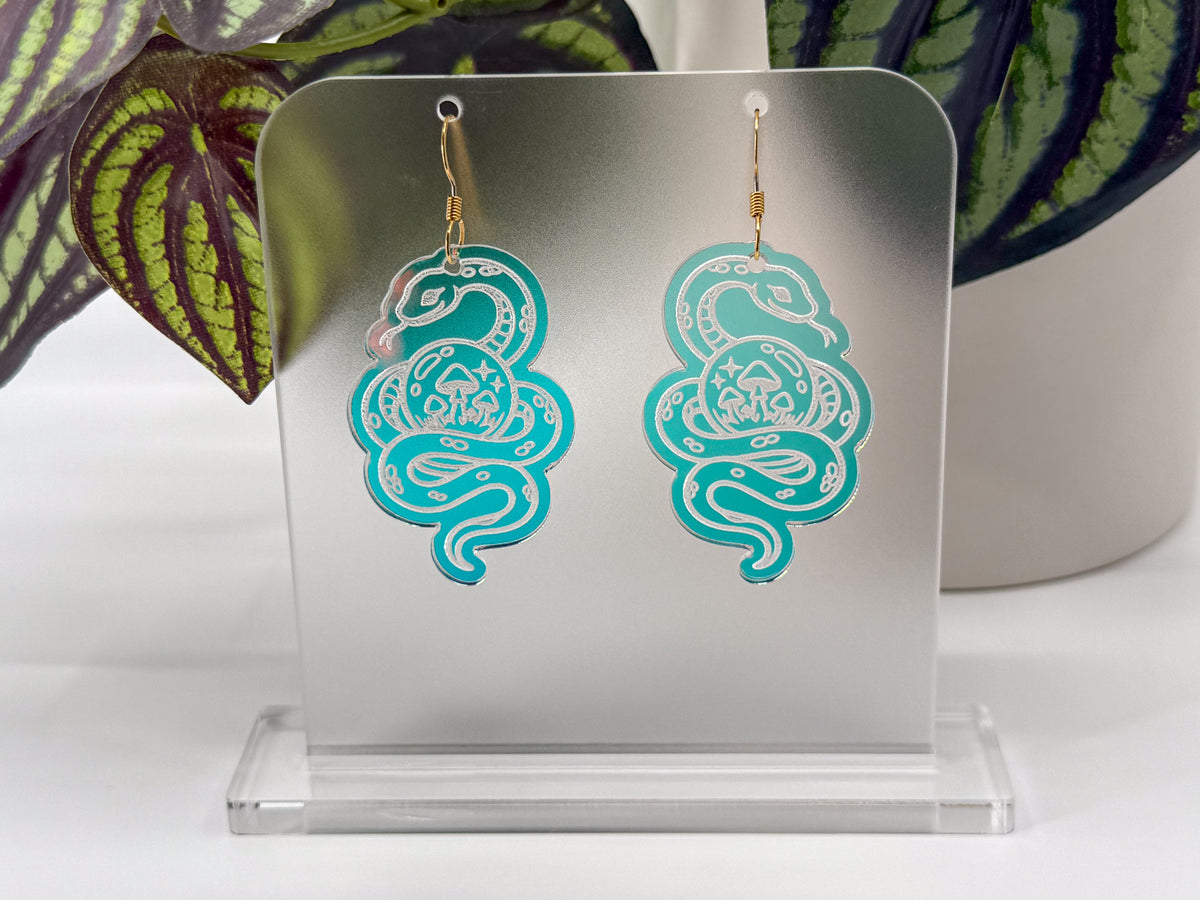 A pair of turquoise and silver snake-shaped dangle earrings displayed on a clear acrylic stand, with green and purple patterned leaves in the background.