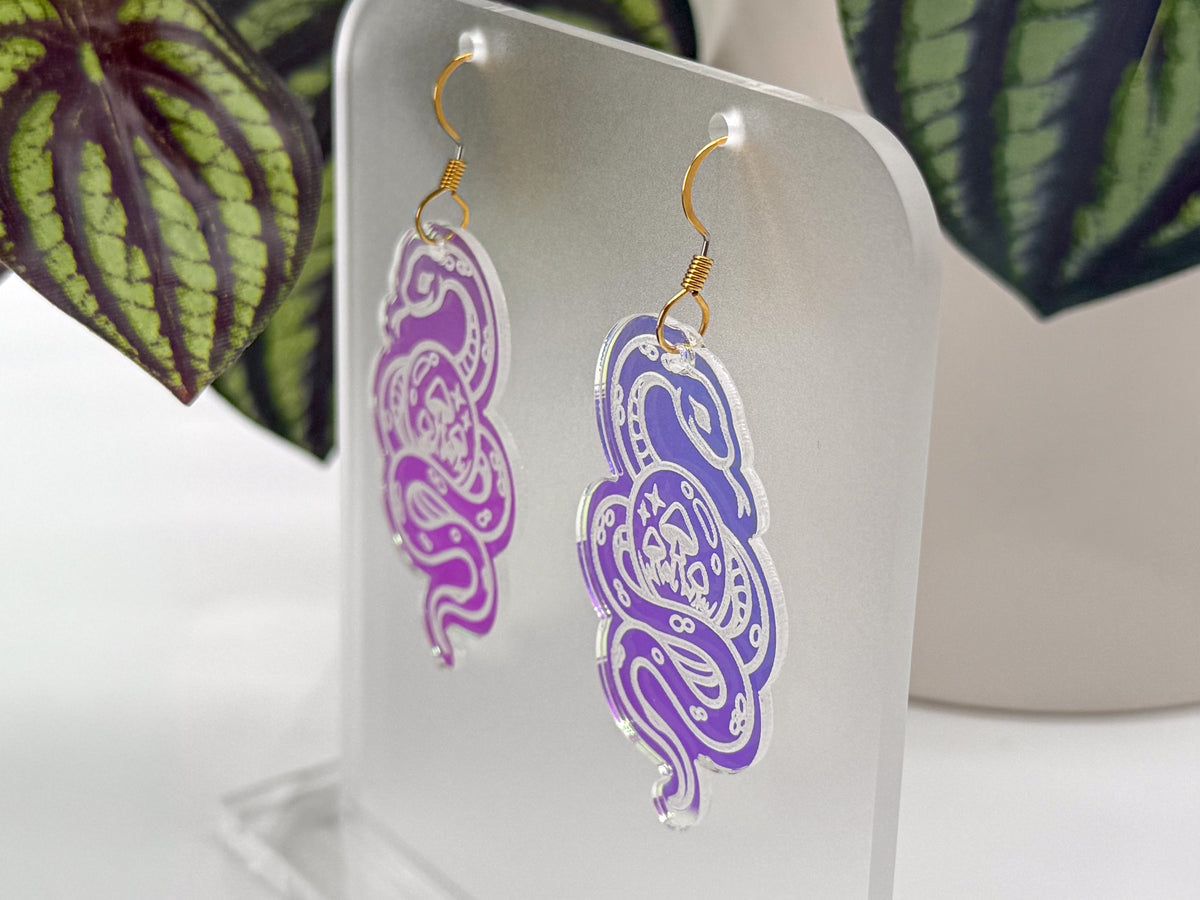 A pair of dangling snake-shaped earrings displayed on a clear acrylic stand. The earrings have a purple-to-pink gradient cutout design with gold fishhook ear wires.