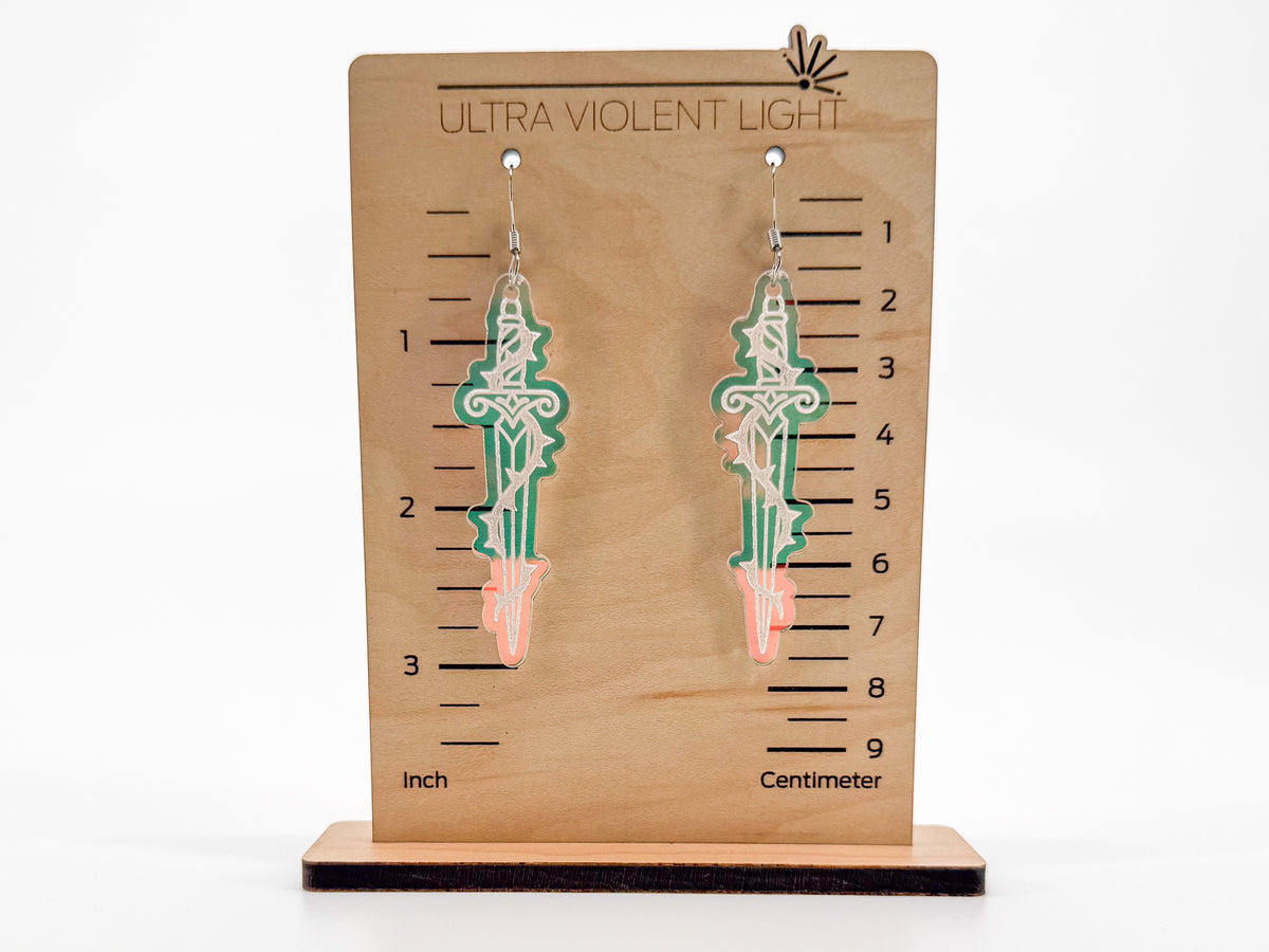 A pair of dangling earrings displayed on a wooden measurement stand labeled 'ULTRA VIOLENT LIGHT' with inch and centimeter markings. The earrings are shaped like swords, engraved on iridescent acrylic, shown against a plain white background.
