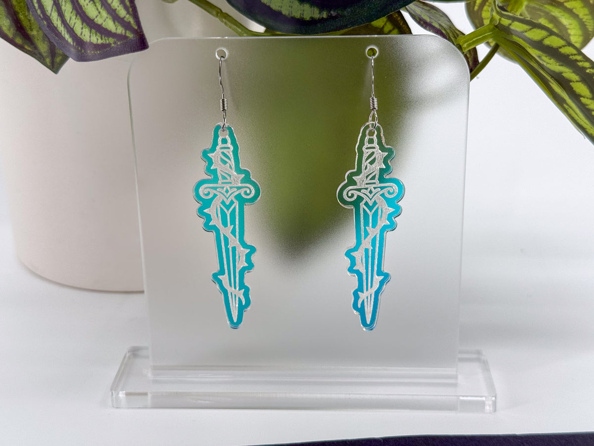 A pair of silver-toned dagger-shaped earrings with turquoise and white cutout detailing, displayed on a clear acrylic stand against a white background with a blurred plant in the background.