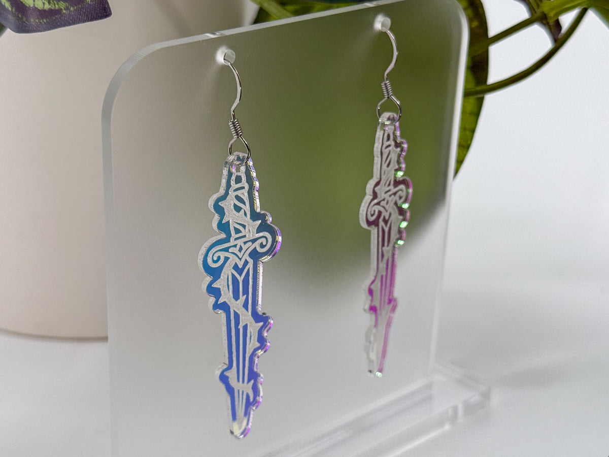 A pair of dangling earrings shaped like ornate swords, displayed on a clear acrylic stand. One earring is iridescent blue to purple, and the other is iridescent pink to purple, both with silver hooks.