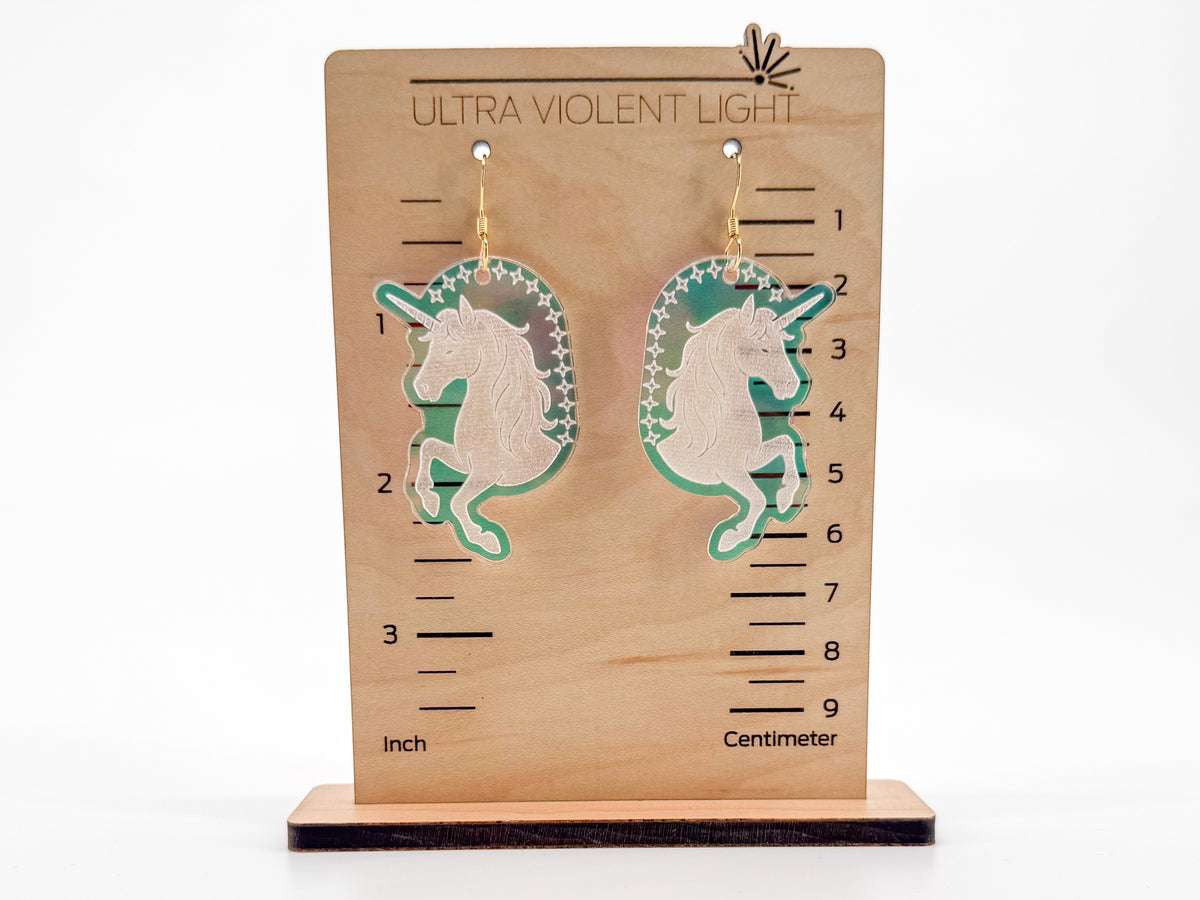 A pair of unicorn-shaped acrylic drop earrings displayed on a wooden measurement stand labeled 'ULTRA VIOLENT LIGHT.' The earrings are made of engraved iridescent acrylic with gold hooks.