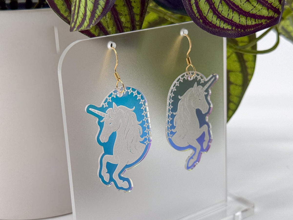 A pair of dangling unicorn-shaped earrings displayed on a clear acrylic stand. The earrings have a holographic finish shifting between blue, purple, and silver, with gold fishhook wires.