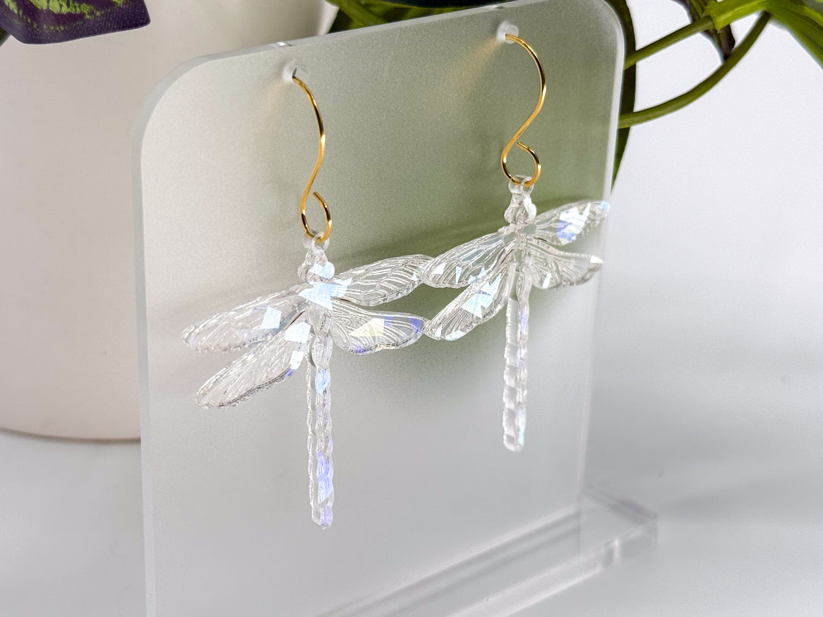 A pair of clear dragonfly-shaped dangling earrings with gold hooks, displayed on a translucent acrylic stand against a soft white background with blurred green leaves.