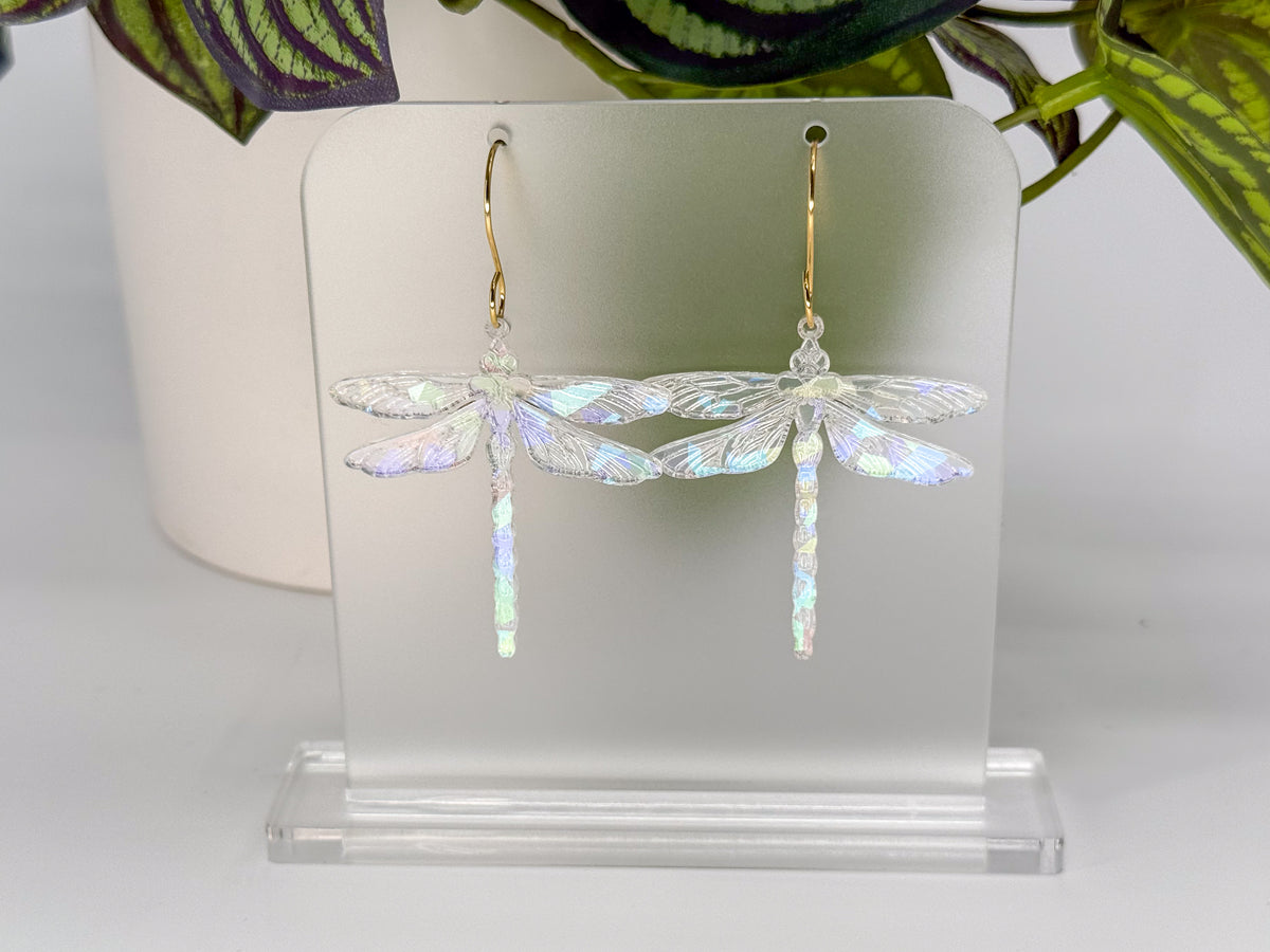 A pair of dragonfly-shaped dangling earrings with iridescent wings and gold hooks, displayed on a clear acrylic stand against a white background with blurred green leaves above.