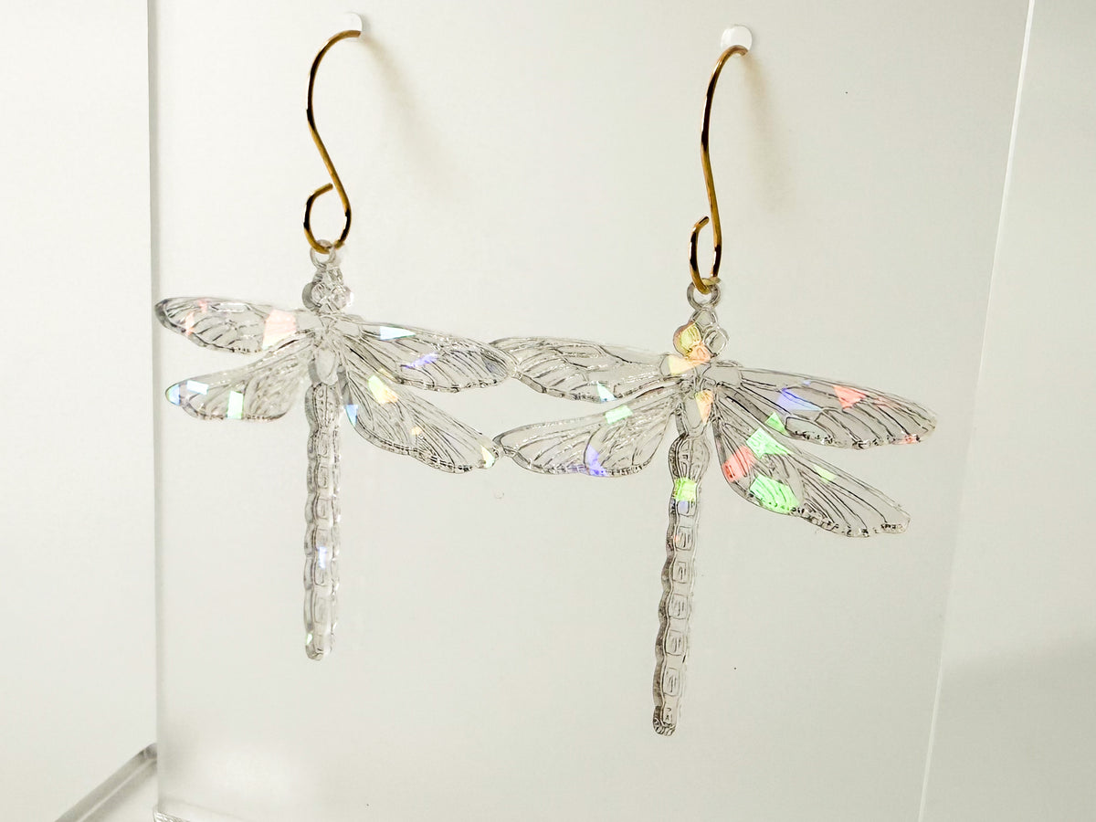 A pair of dragonfly-shaped dangling earrings made of clear faceted material with iridescent wing details and gold-tone hooks, shown against a plain white background.