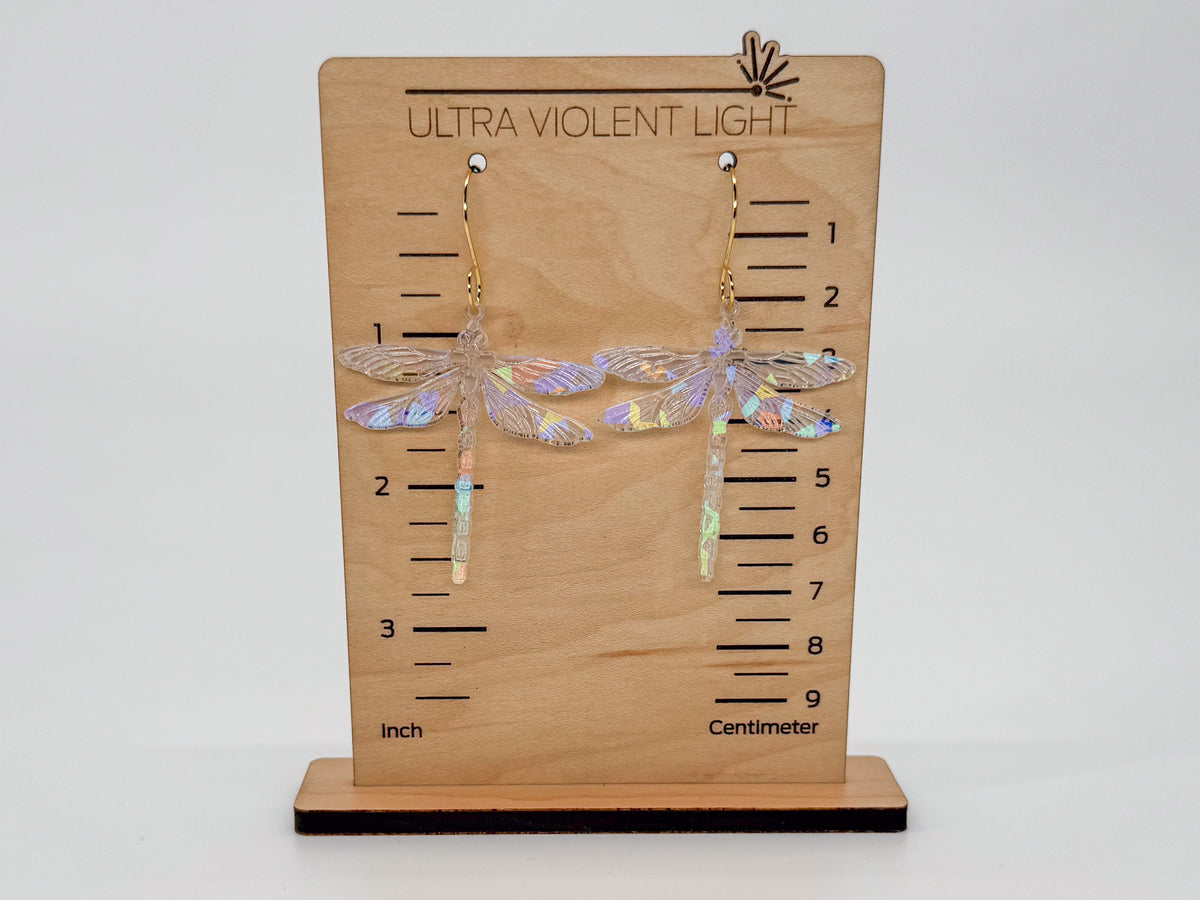 A pair of dragonfly-shaped dangling earrings displayed on a wooden measurement stand labeled 'ULTRA VIOLENT LIGHT.' The earrings have iridescent clear wings and gold hooks.
