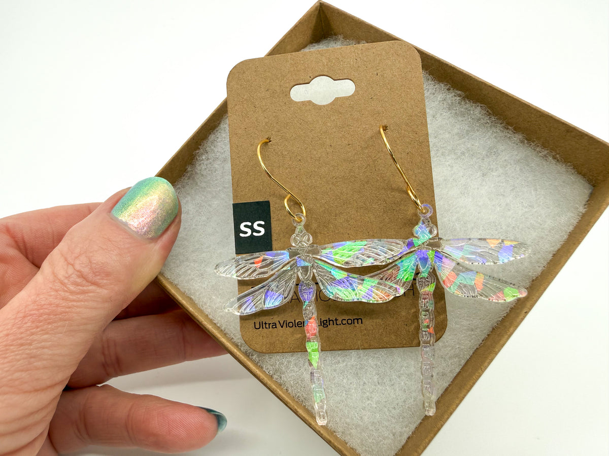 A hand with iridescent nail polish holds a pair of dangling dragonfly earrings on a brown display card. The earrings have iridescent holographic wings and gold hooks, presented in a brown box with white padding.