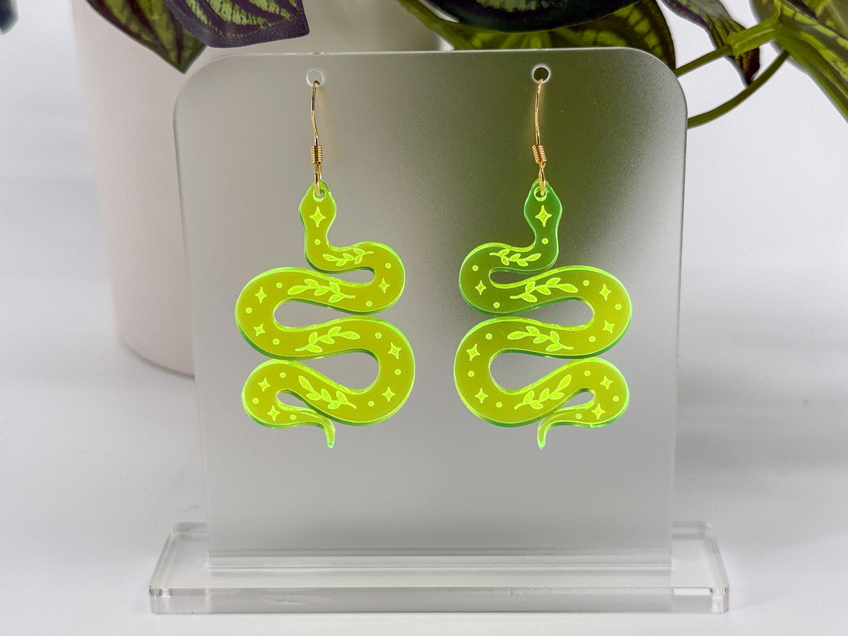 A pair of neon yellow-green snake-shaped dangle earrings with gold hooks, displayed on a clear acrylic stand against a white background with a blurred plant in the background.