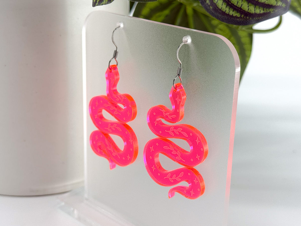A pair of neon pink snake-shaped dangle earrings with silver fishhook wires, displayed on a clear acrylic stand.