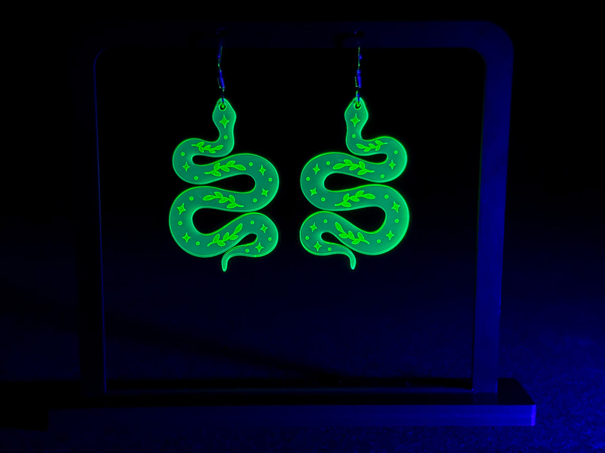 Two neon green glowing snake-shaped earrings hanging from ear hooks, displayed against a dark black background with a blue UV light effect.