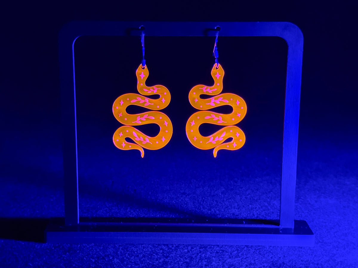 A pair of snake-shaped dangling earrings displayed on a black stand under blue light. The earrings are bright orange and pink.