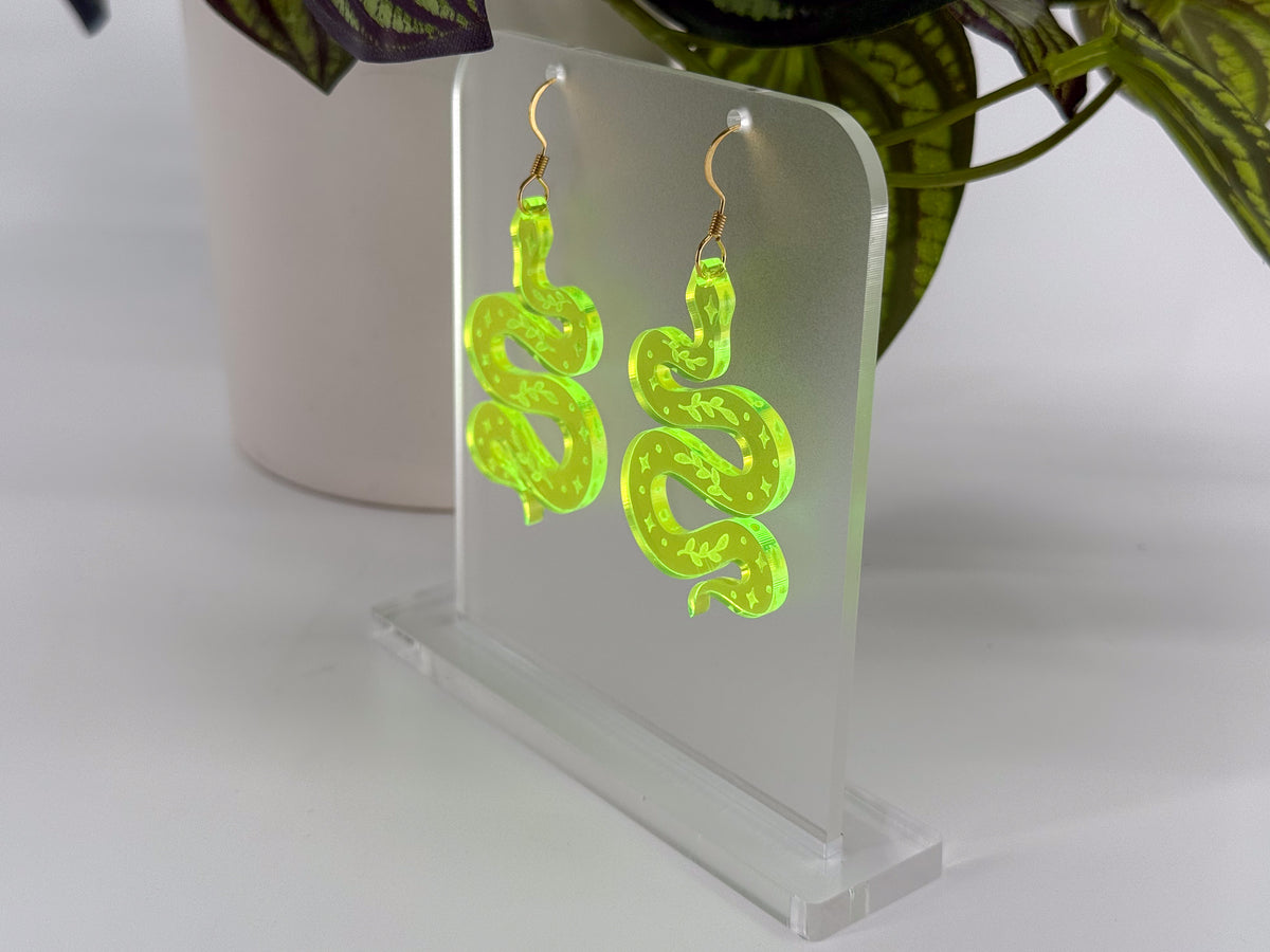 A pair of neon green snake-shaped dangle earrings displayed on a clear acrylic stand against a white background with a blurred potted plant.