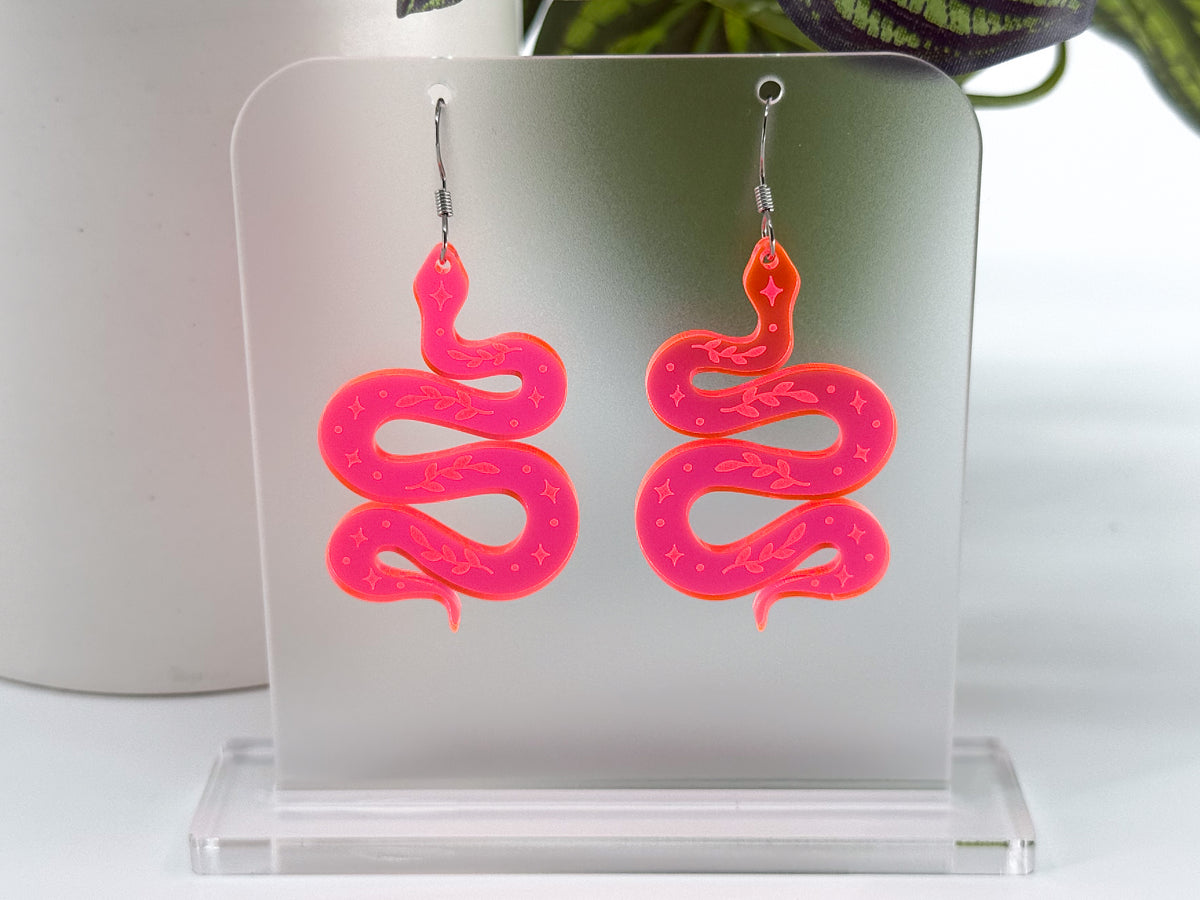 A pair of fluorescent pink snake-shaped dangle earrings displayed on a clear acrylic stand against a bright white background with a blurred plant in the corner.