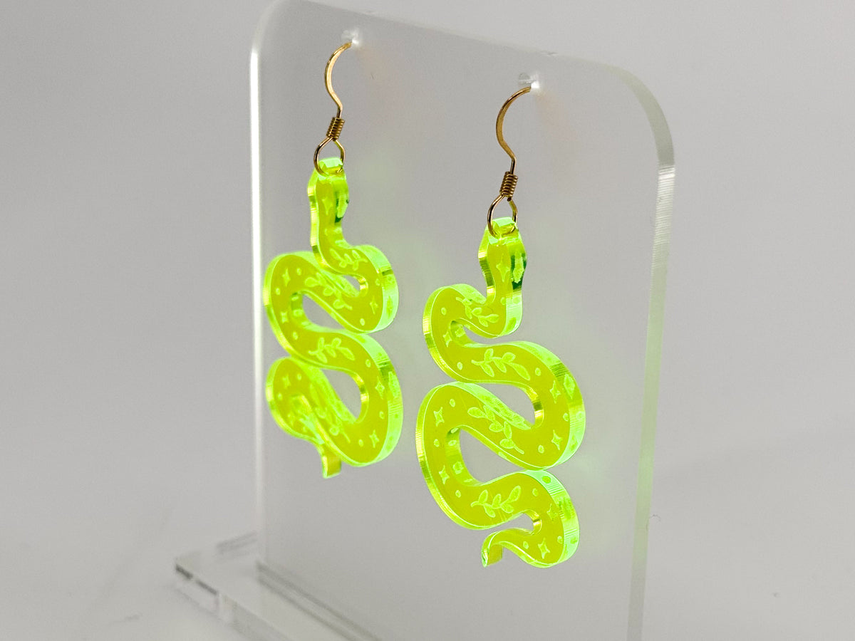 A pair of neon yellow-green snake-shaped dangle earrings with gold fishhook wires, displayed on a clear acrylic stand against a white background.