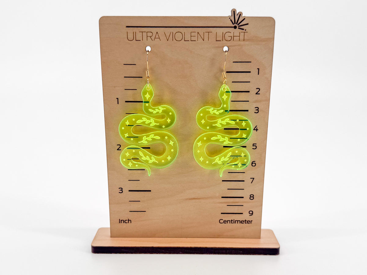 A pair of fluorescent green snake-shaped dangle earrings displayed on a wooden height-measuring stand labeled 'ULTRA VIOLENT LIGHT' with inch and centimeter markings, against a plain white background.