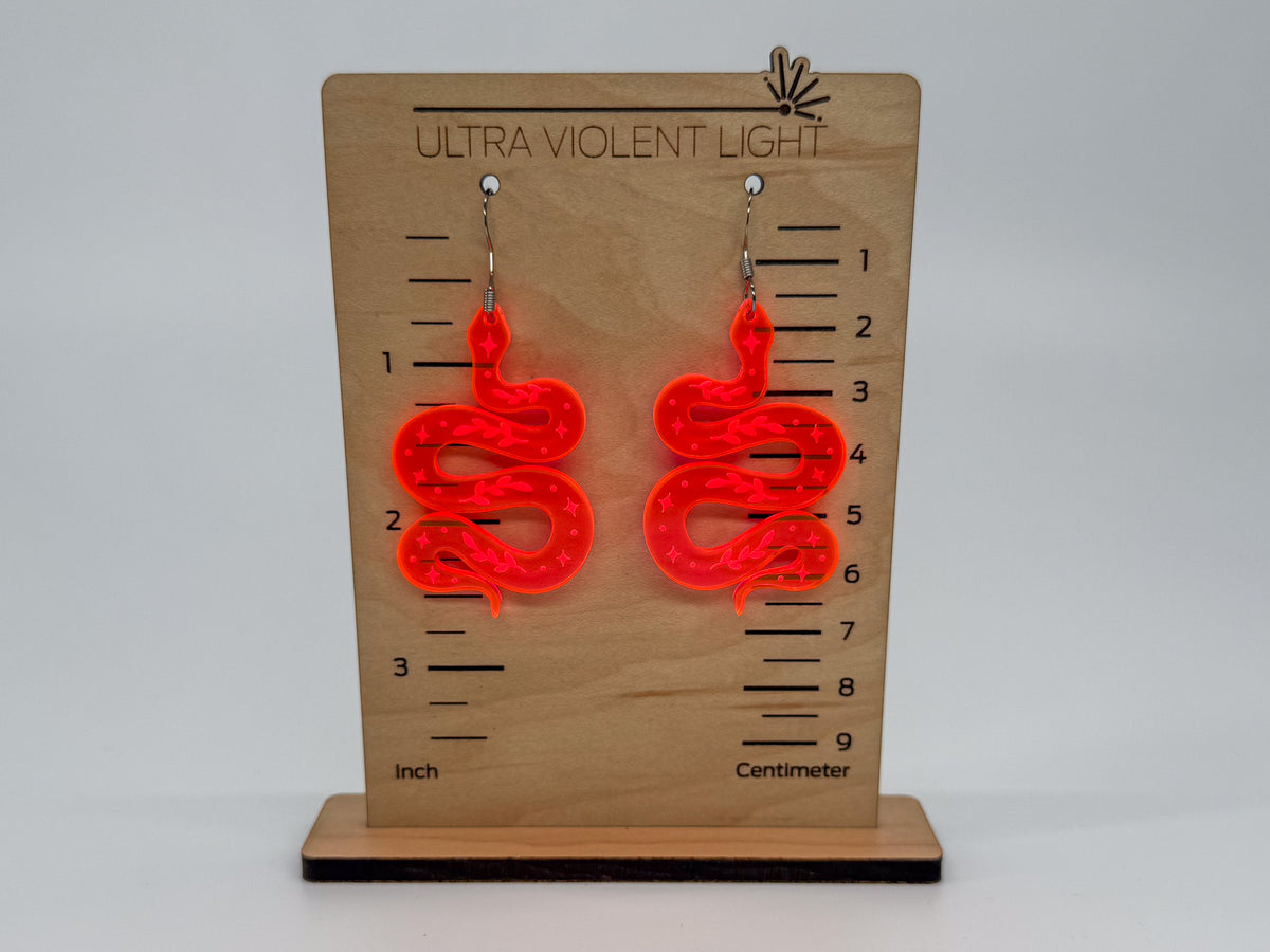 A pair of bright neon orange-red snake-shaped dangle earrings displayed on a wooden measurement stand labeled 'ULTRA VIOLENT LIGHT' with inch and centimeter markings.