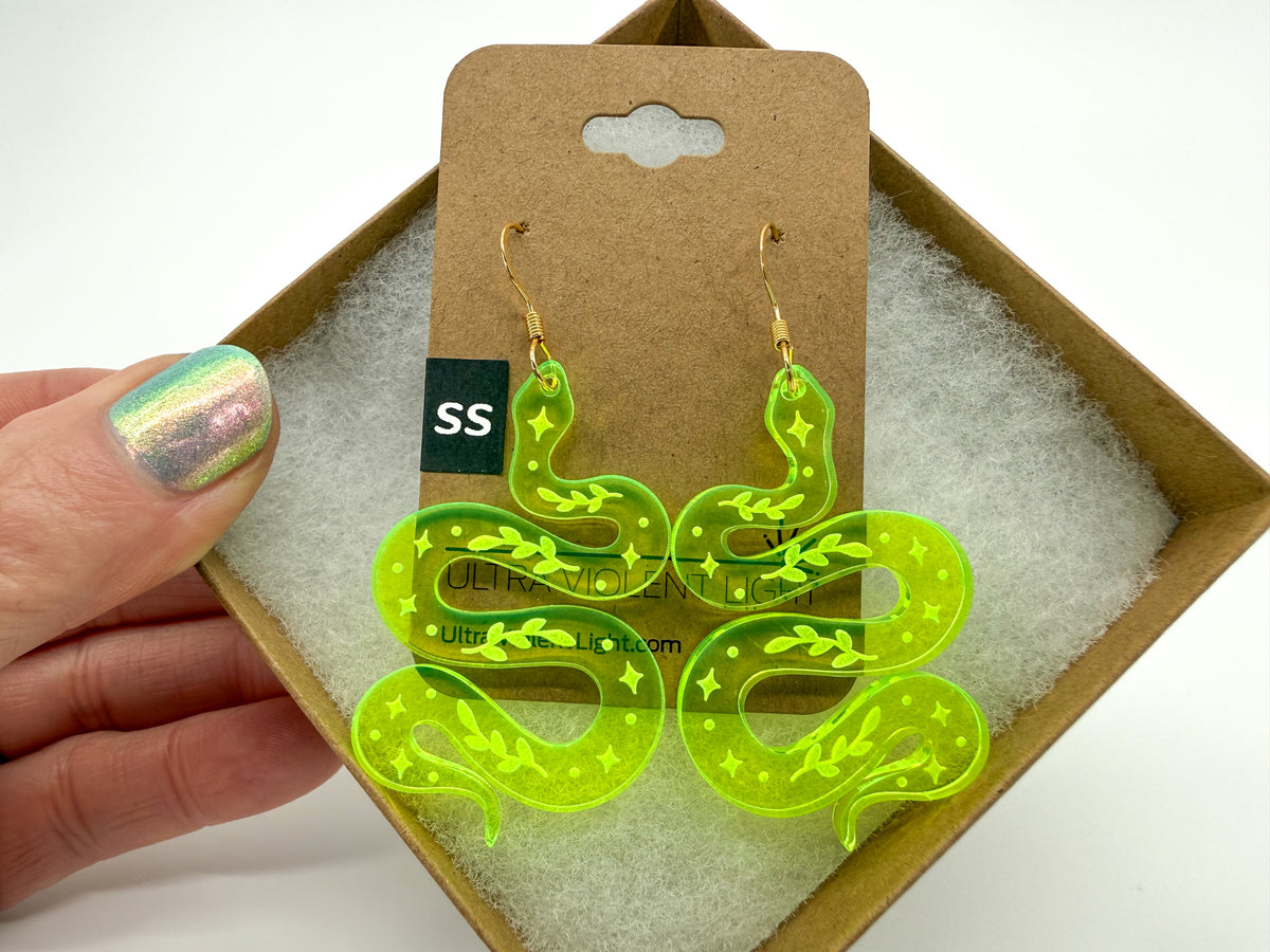 A hand with iridescent nail polish holds a pair of neon yellow-green snake-shaped drop earrings with gold hooks, displayed on a brown cardboard backing card in front of an open gift box with white padding.