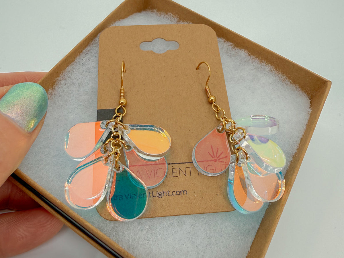 A pair of dangling earrings displayed on a brown card insert inside a kraft gift box. The earrings have gold-tone hooks and multiple translucent acrylic teardrop shapes in iridescent colors.