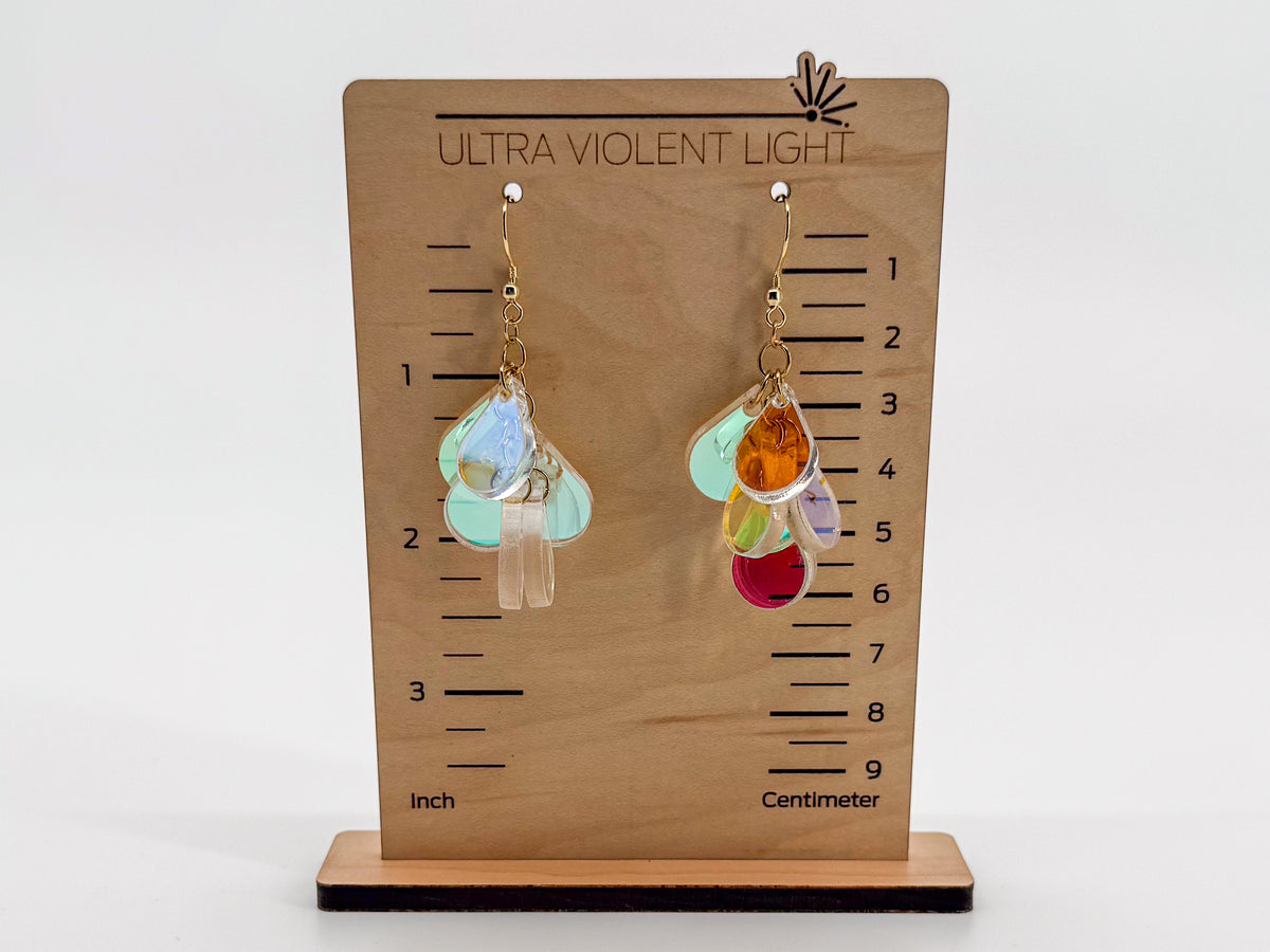 A pair of dangling earrings displayed on a wooden measurement stand labeled 'ULTRA VIOLENT LIGHT.' The earrings have gold-tone hooks and multiple iridescent translucent drops in pale blue, mint, aqua, peach, orange, and red.