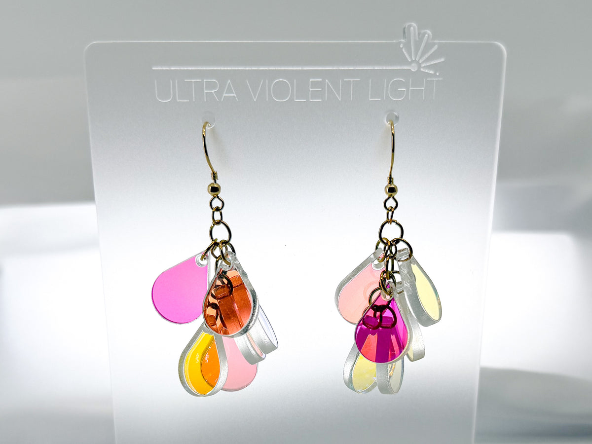 A pair of dangling earrings displayed on a clear acrylic stand labeled 'ULTRA VIOLENT LIGHT.' The earrings have gold-tone hooks and multiple translucent petal-like charms in pink, orange, yellow, and clear/white.