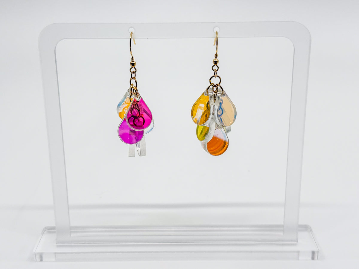 A pair of dangling earrings displayed on a clear acrylic stand against a white background. Each earring has a gold-tone hook and multiple translucent teardrop-shaped pieces in pink, orange, yellow, and clear.