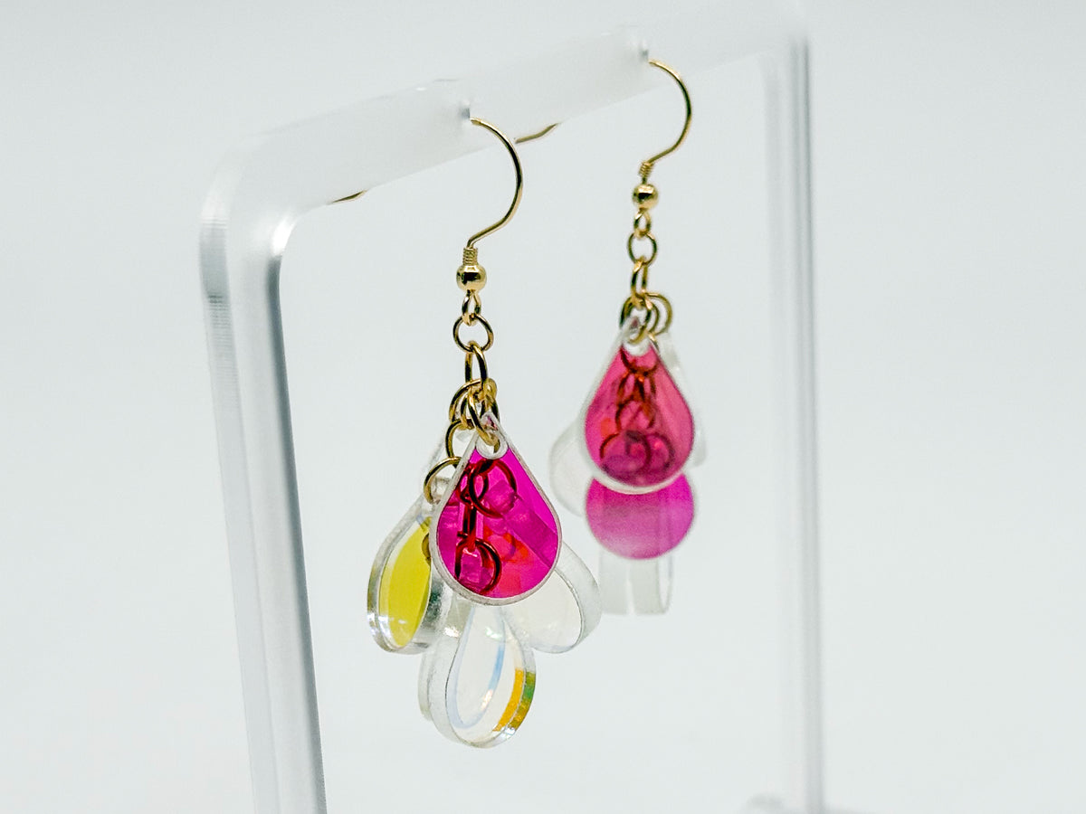 A pair of dangling earrings displayed on a clear acrylic stand against a white background. Each earring has a gold fishhook wire, a small gold ring, and a cluster of teardrop-shaped clear acrylic drops.
