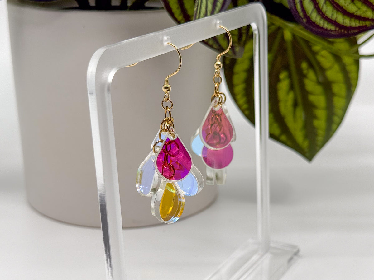 A pair of dangling earrings displayed on a clear acrylic stand. Each earring has a gold fishhook wire and a cluster of teardrop-shaped translucent charms in pink, light blue, and yellow.
