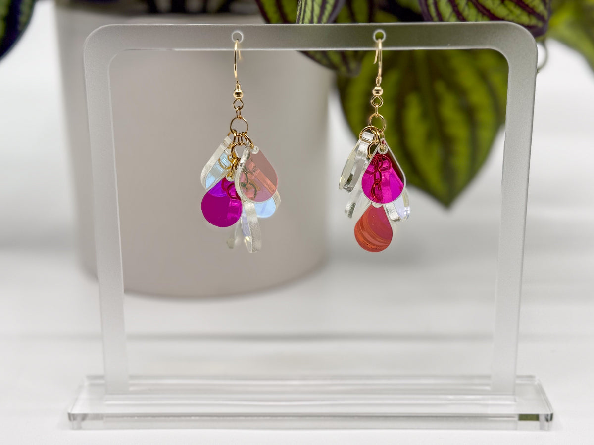 A pair of dangling earrings displayed on a clear acrylic stand. Each earring has a gold fishhook ear wire with a cluster of dangling iridescent teardrop shaped charms.