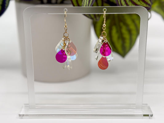 A pair of dangling earrings displayed on a clear acrylic stand. Each earring has a gold fishhook ear wire with a cluster of dangling iridescent teardrop shaped charms.