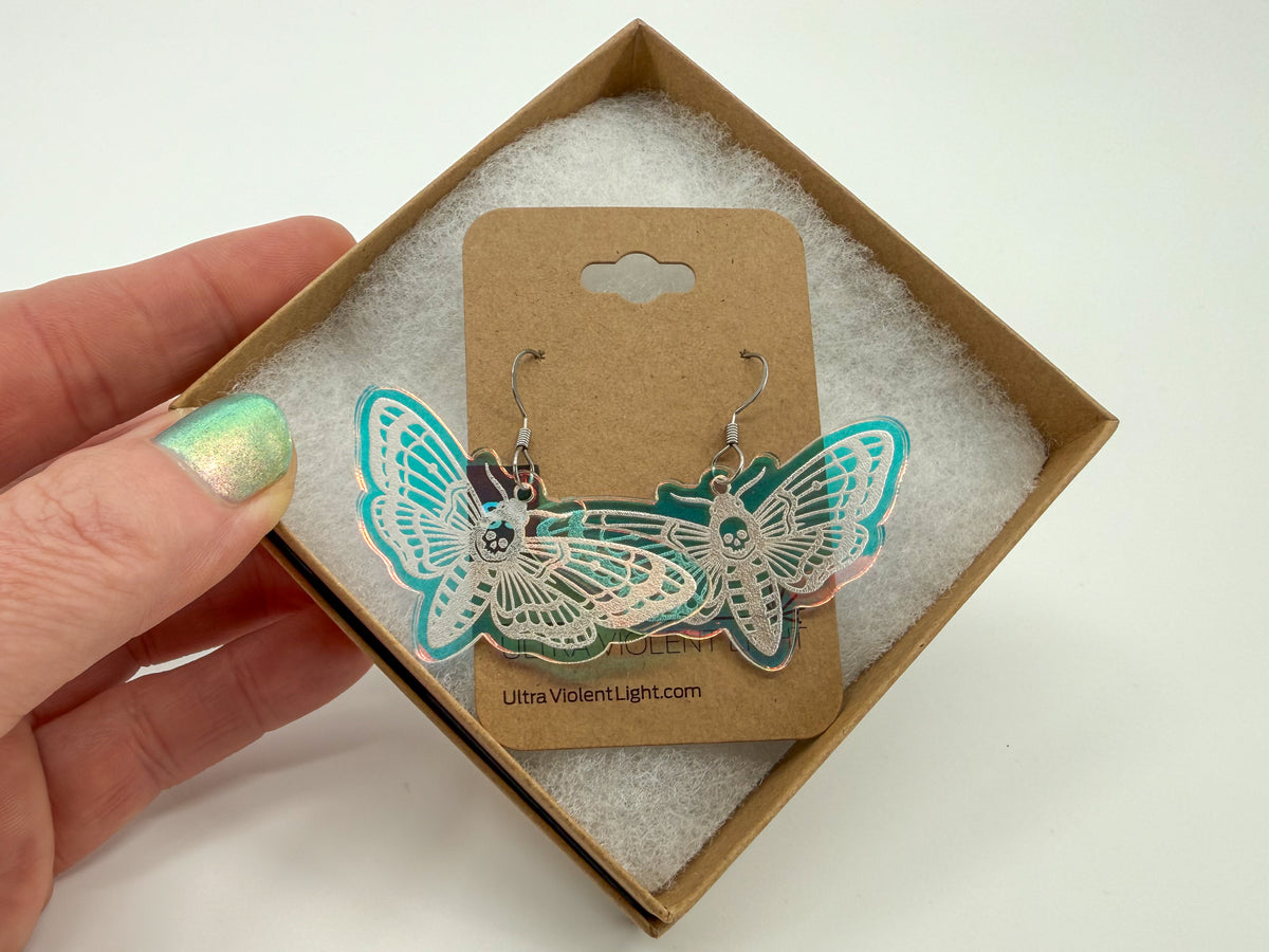 A hand with iridescent green nail polish holds an open square kraft paper box lined with white padding, displaying a pair of moth-shaped dangle earrings on a brown card. The earrings are engraved from iridescent acrylic, appearing turquoise here.
