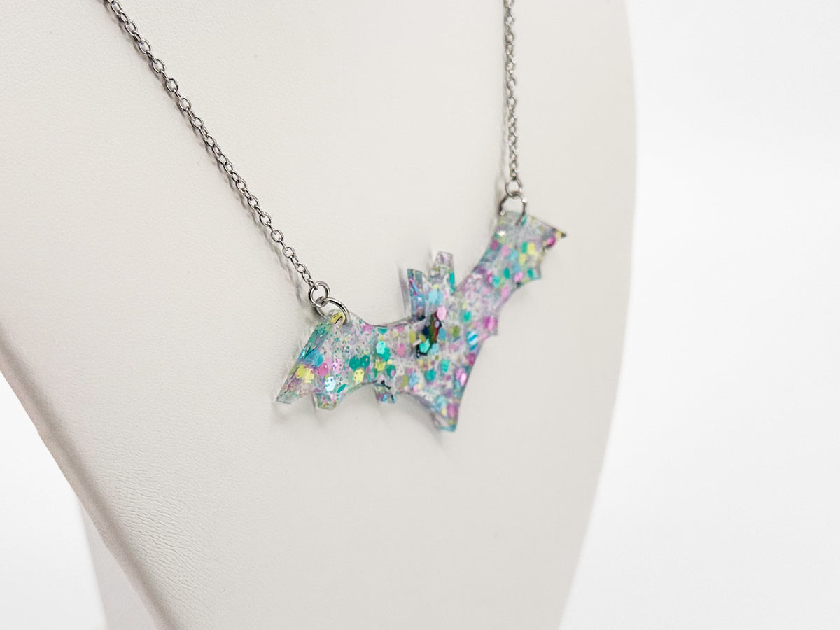 Angled close-up of a clear acrylic bat necklace with pastel confetti and small reflective bats on a white display bust.