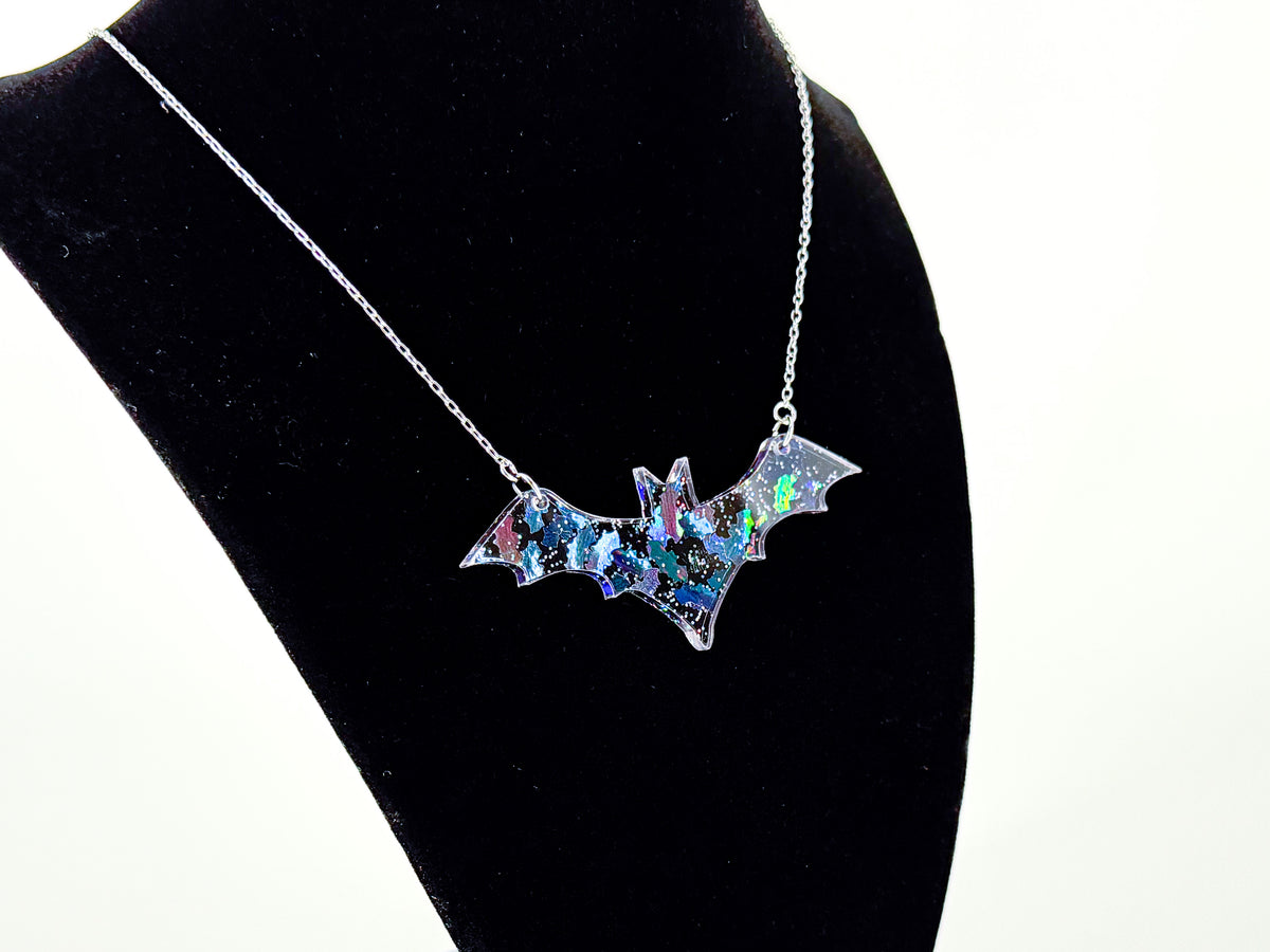Iridescent bat shapes reflecting blue, green and pink inside an acrylic bat pendant on a black velvet display bust.