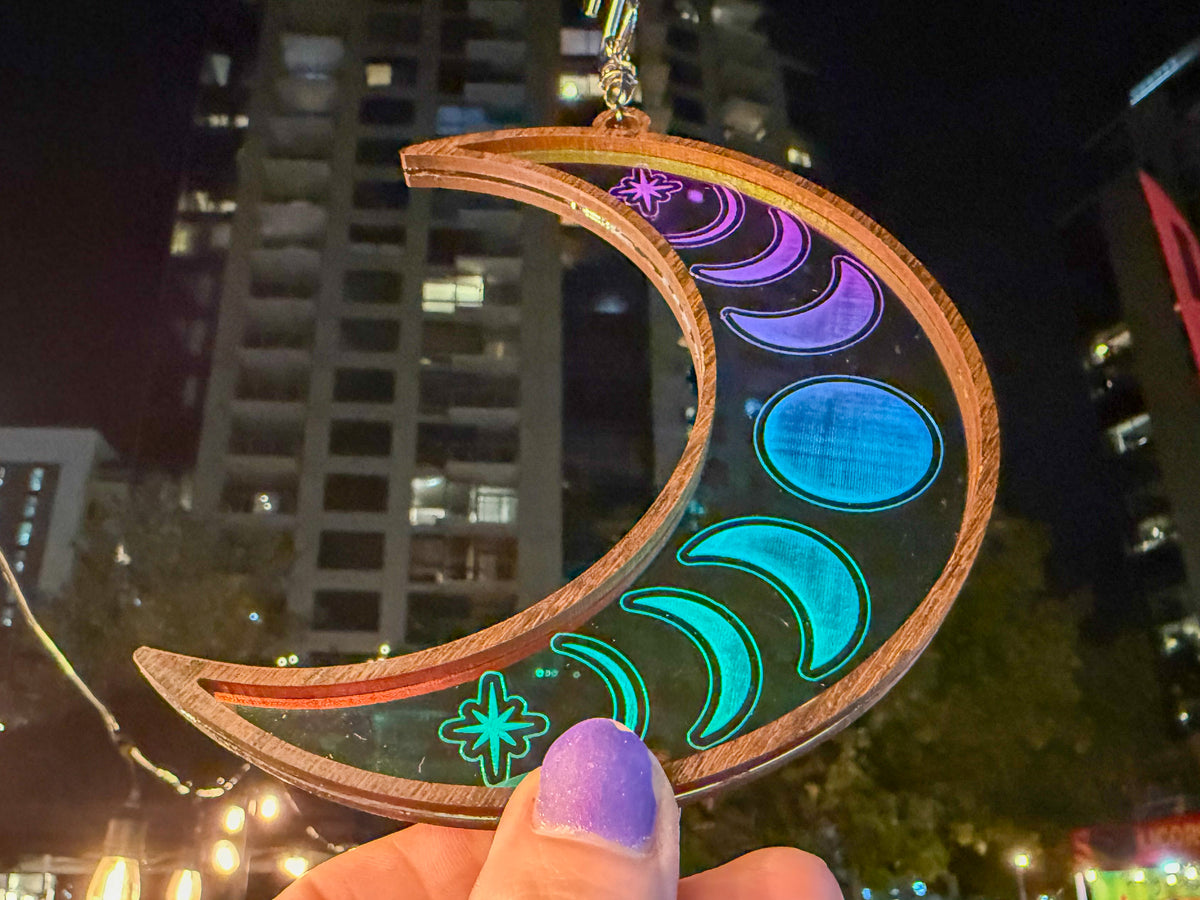 Celestial moon suncatcher with a wood frame and engraved moon phases reflecting purple, blue, and green, held against a city backdrop.