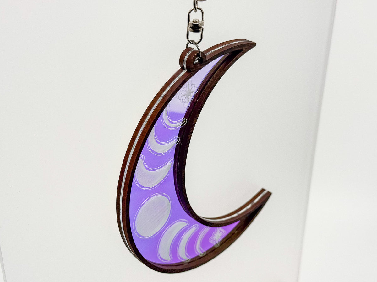 Angled view of a wood-framed crescent moon suncatcher with a purple iridescent center engraved with moon phases and stars.