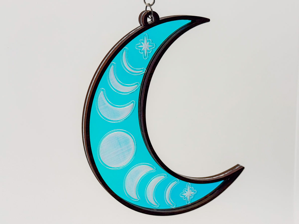 Celestial moon suncatcher with a bright turquoise iridescent center, engraved moon phases and stars, and a dark wood crescent frame.