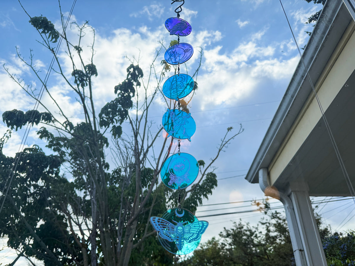 Full-length whimsigothic acrylic mobile with six linked engraved charms reflecting blue and teal against the sky.