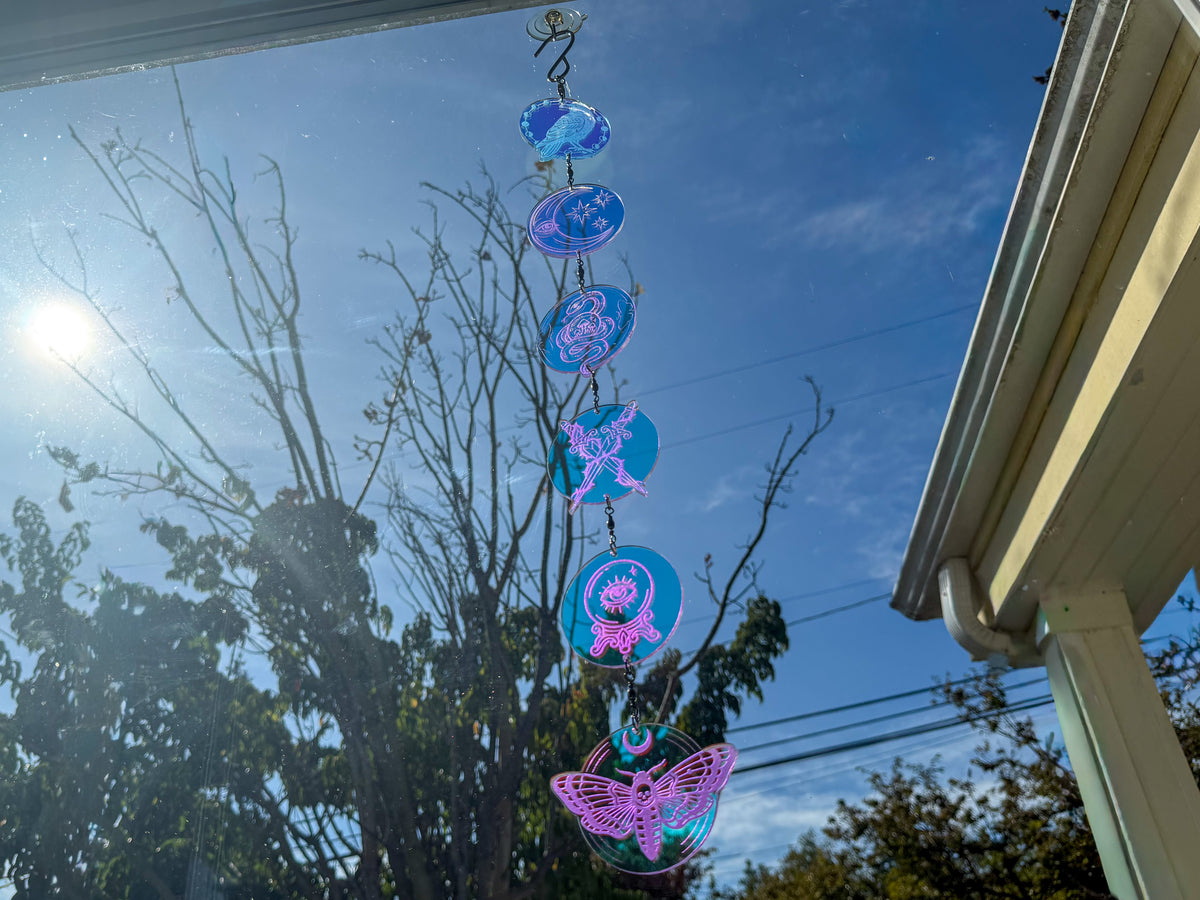 Whimsigothic suncatcher with raven, moon, snake, crossed daggers, crystal ball, and moth charms reflecting pink and blue.