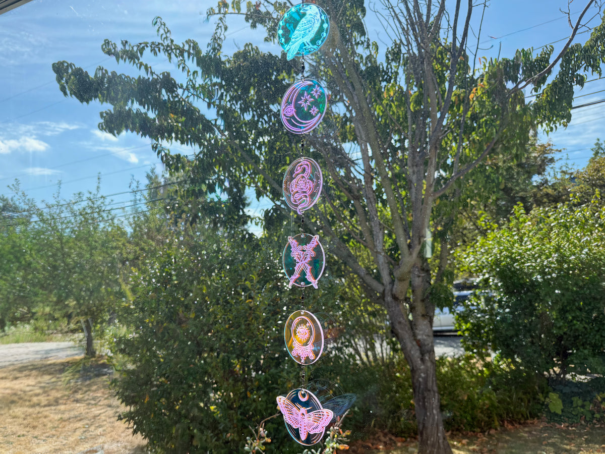 Iridescent engraved suncatcher hanging against garden greenery, with six charms reflecting teal, pink, and gold.