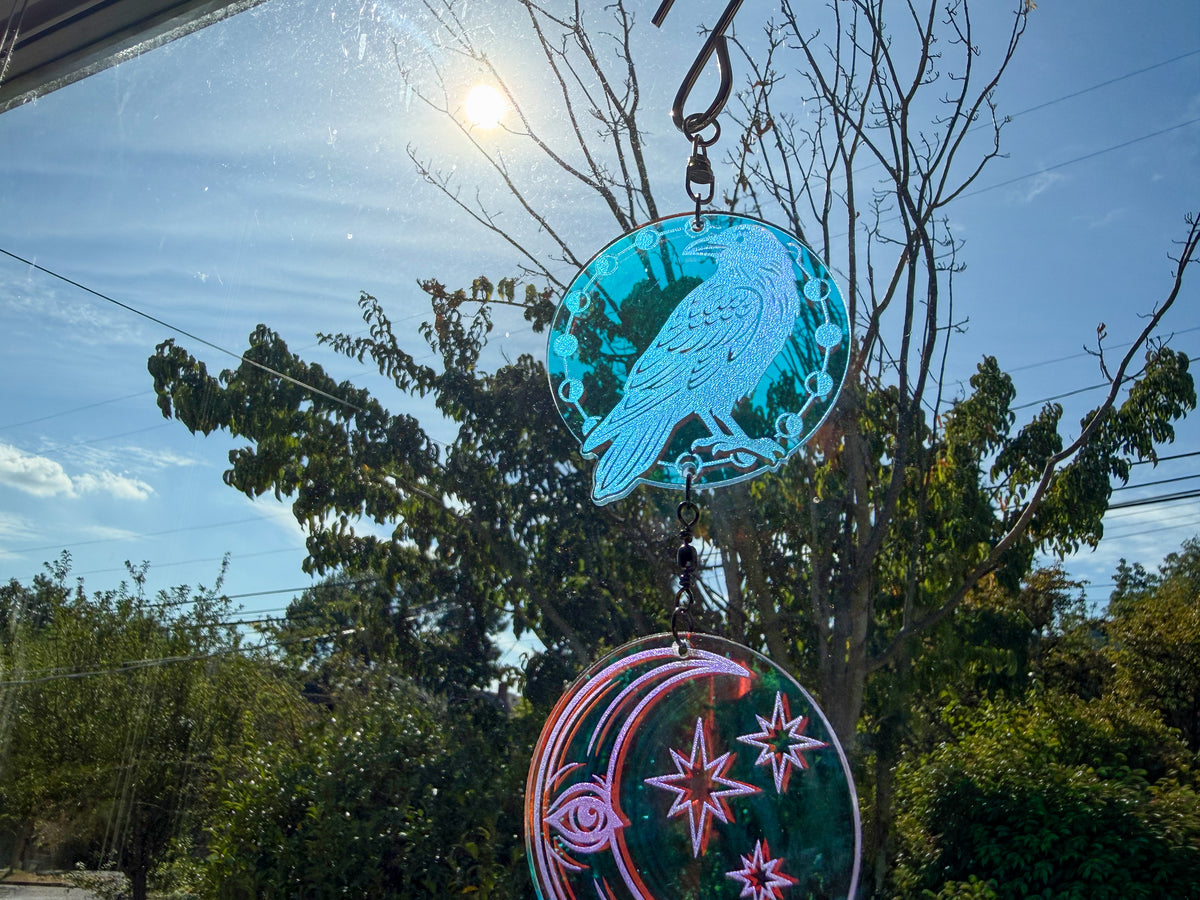 Close-up of an engraved raven charm above a crescent moon and stars, reflecting teal and pink in sunlight.