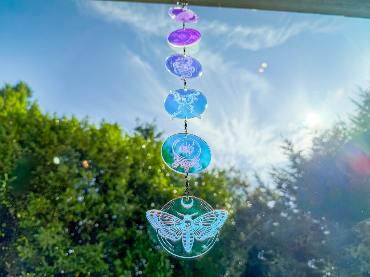 Iridescent whimsigothic suncatcher hanging in a sunny window, with engraved charms glowing purple, blue, and teal above a moth.