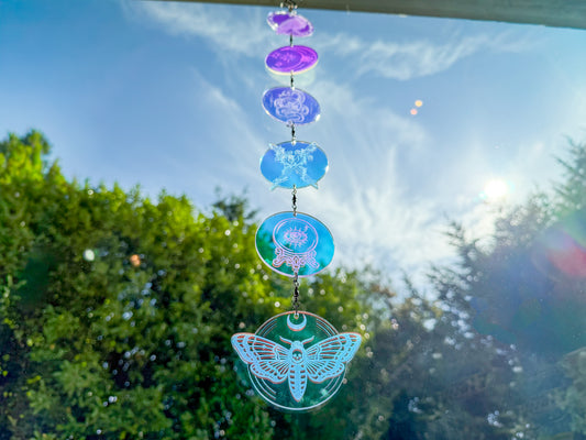 Iridescent whimsigothic suncatcher hanging in a sunny window, with engraved charms glowing purple, blue, and teal above a moth.