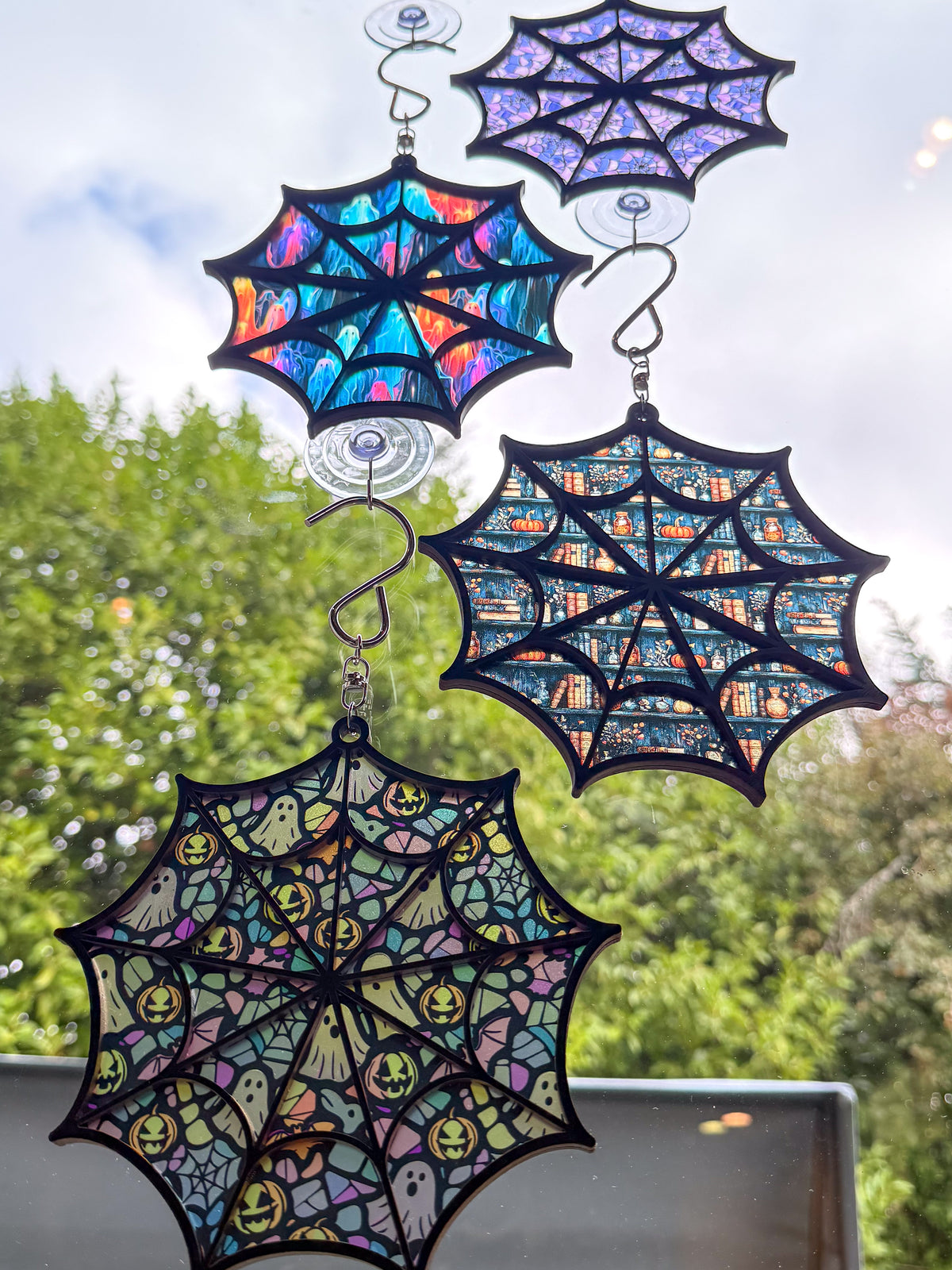 Four black-framed spiderweb suncatchers hanging in a sunny window, showing purple webs, colorful ghosts, fall bookshelves, and whimsical Halloween designs.