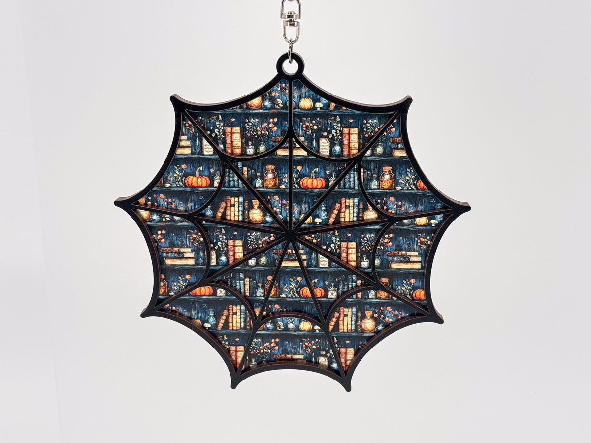 Front view of a black-framed spiderweb suncatcher with a bookshelf pattern featuring books, orange pumpkins, bottles, and flowers.